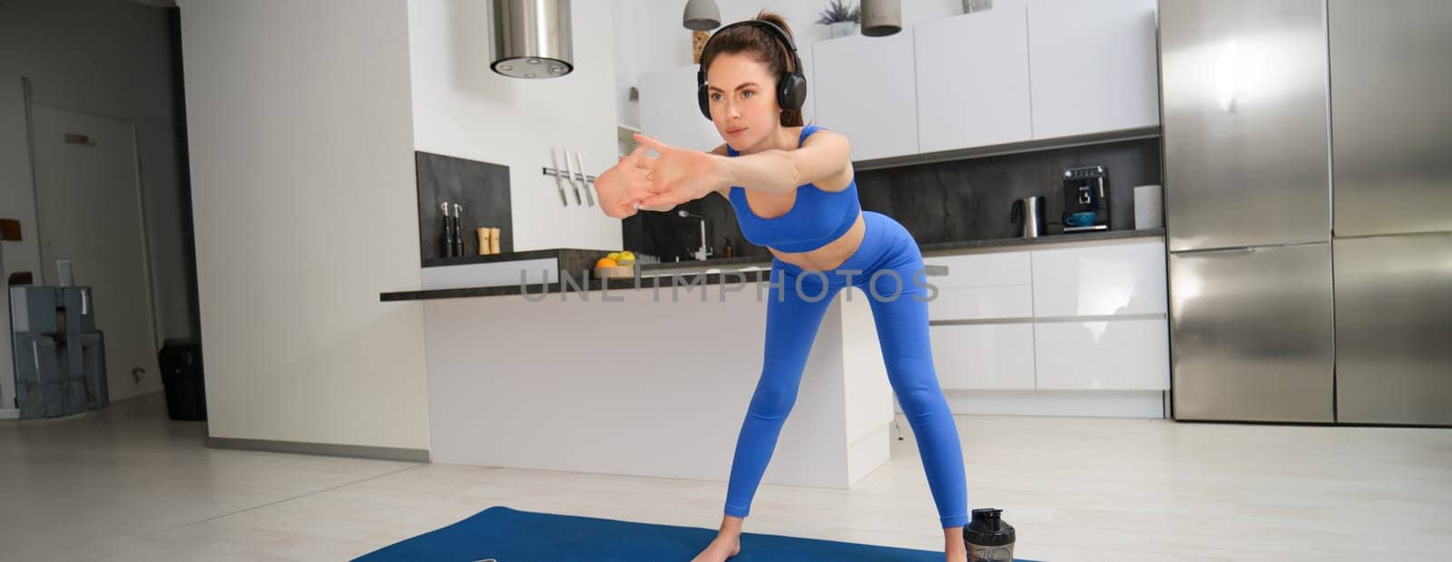 Portrait of young woman doing aerobics exercises in living room, sport training at home, standing on rubber fitness mat and doing workout.