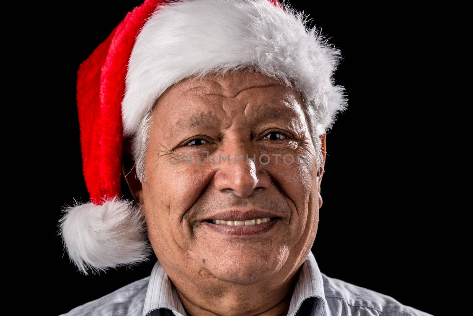 Aged gentleman,wearing a red and white Old Father Christmas hat,is smiling. Bright teeth are showing. Black background. Focus on eyes. Ample depth of field. Seasonal theme. Older gentleman wearing a red and white Santa hat,smiles. He shows his white teeth. Black background. Focused on the eyes. Appropriate depth of field. Theme Christmas.