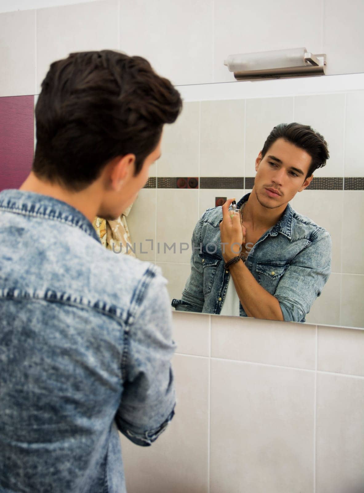 Attractive young man looking at bathroom mirror, spraying cologne or perfume