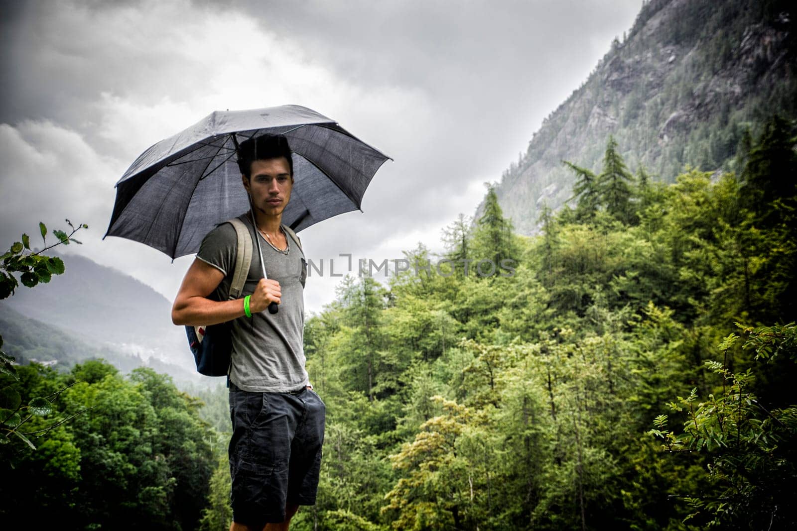 Smiling handsome young man holding an umbrella on a stormy day as he takes a walk along a trail in the mountains, looking at camera
