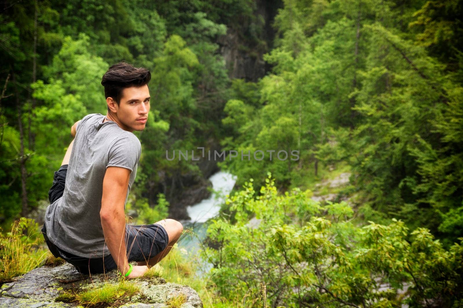 Handsome muscular young man sitting in lush green mountain scenery looking at camera
