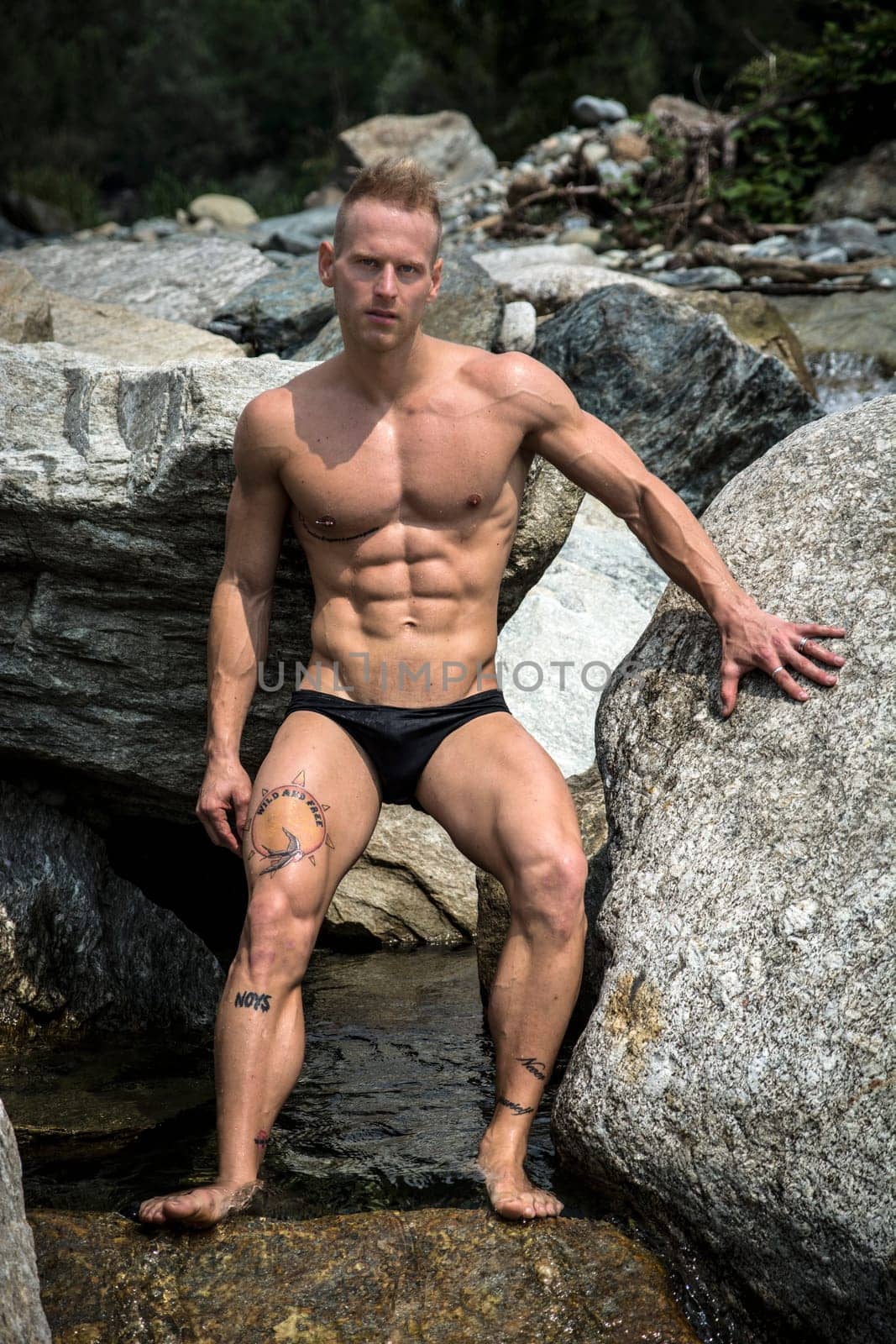 Handsome young muscle man in water pond against rock, naked wearing only bathing suit