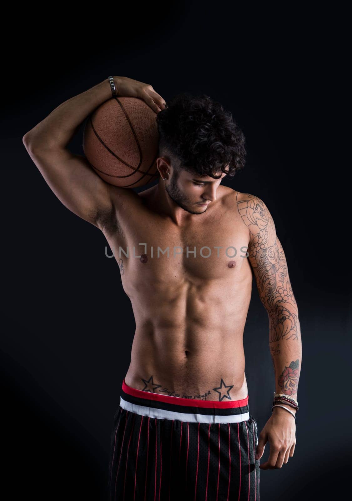 Young athletic man on dark background holding basketball ball on his shoulder, looking down