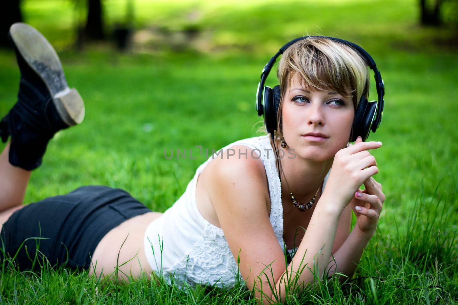 Beautiful young girl listening to music on her headphones as she relaxes in the garden or park lying on her stomach on the green grass