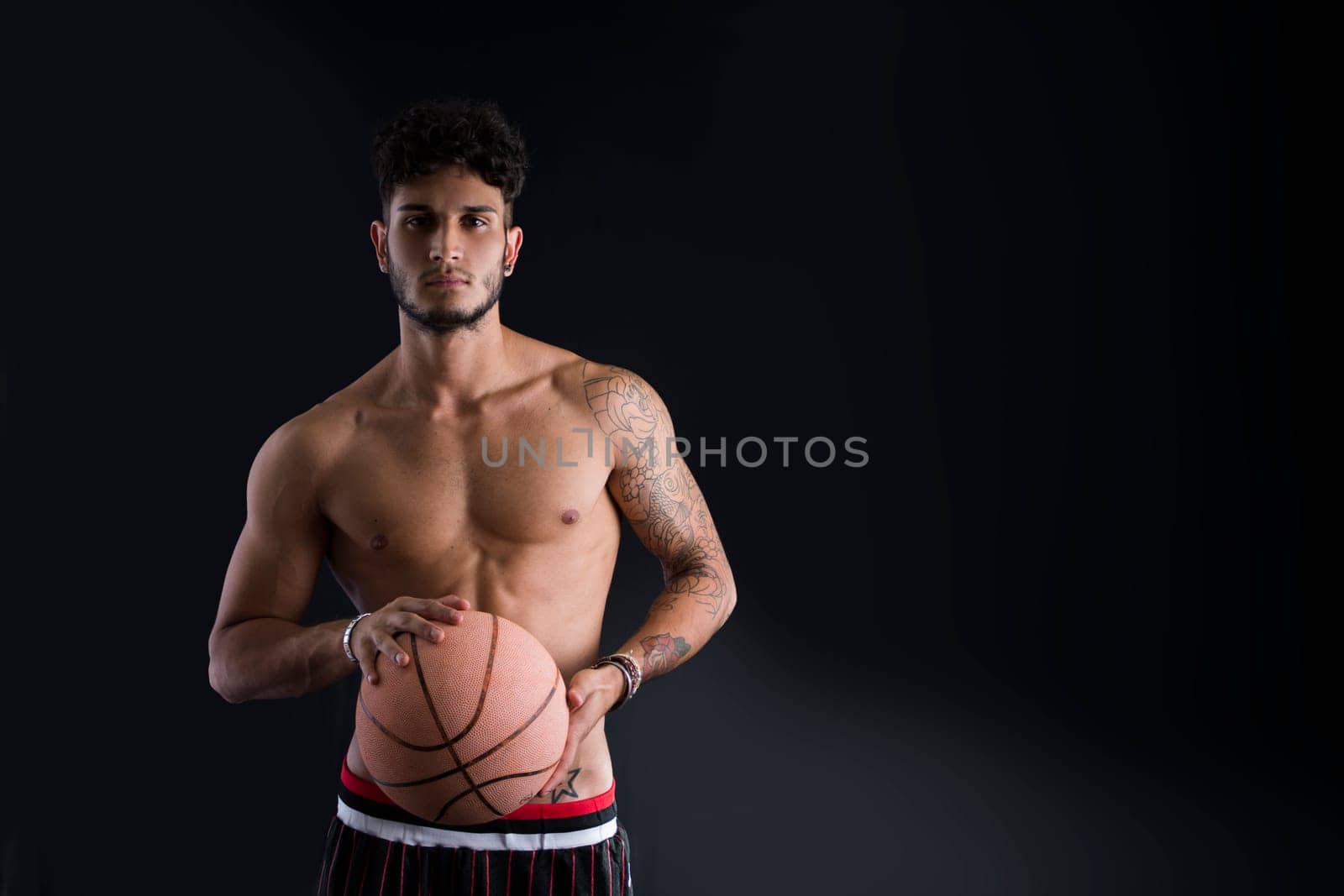 Young athletic man on dark background holding basketball ball, looking at camera