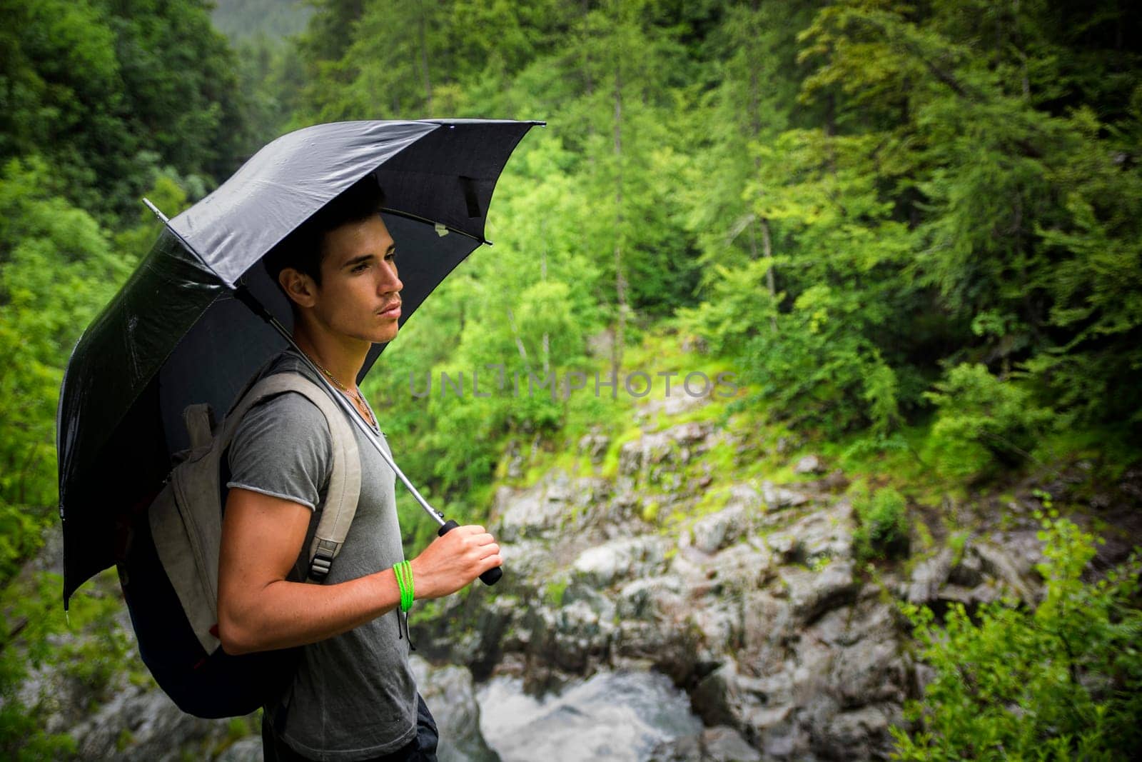 Smiling handsome young man holding an umbrella on a stormy day as he takes a walk along a trail in the mountains, looking far away