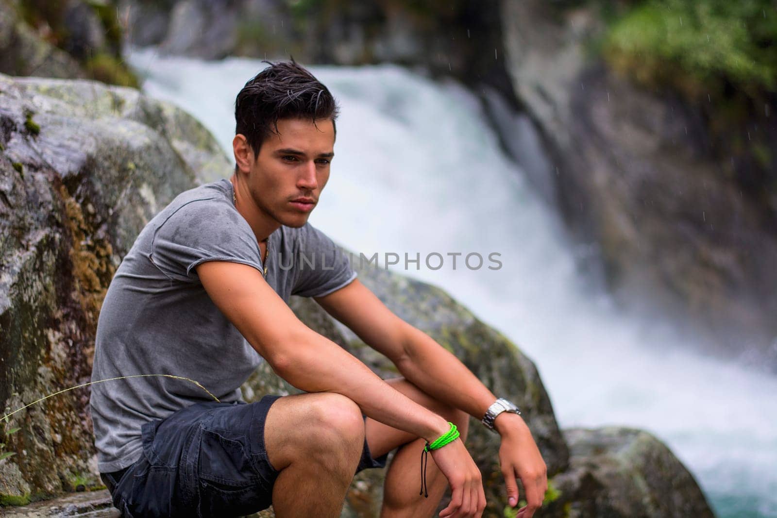 Handsome young man near mountain waterfall sitting on rocks, looking down
