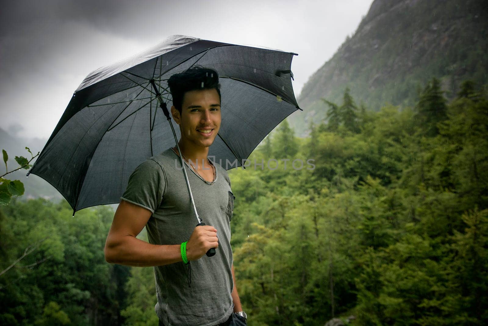 Smiling handsome young man holding an umbrella on a stormy day as he takes a walk along a trail in the mountains turning to smile at the camera