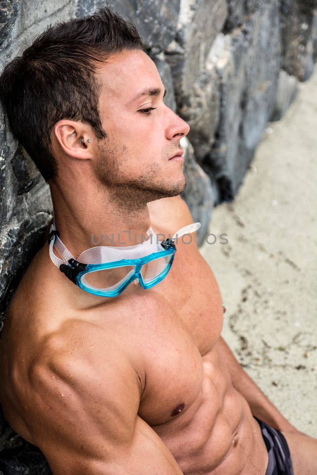 Handsome muscular fit young man relaxing at the beach sitting on the sand leaning against a wall with his swimming goggles around his neck, closeup portrait in profile