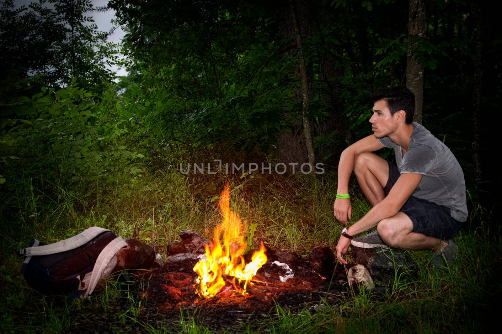 Young man camping in the mountains crouching alongside a burning campfire with his rucksack staring thoughtfully into the distance, profile view