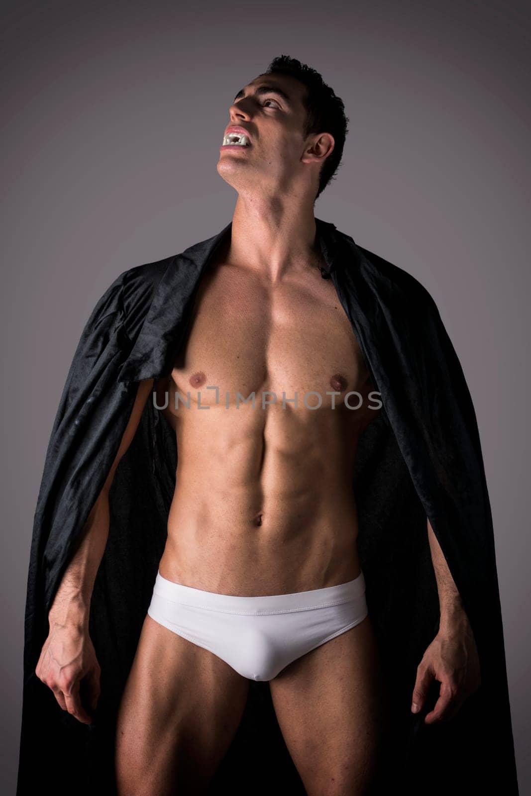 Naked muscular fit young man in briefs posing as a vampire or Dracula in a black cloak showing off his powerful body rasing his head to the sky bearing his fangs