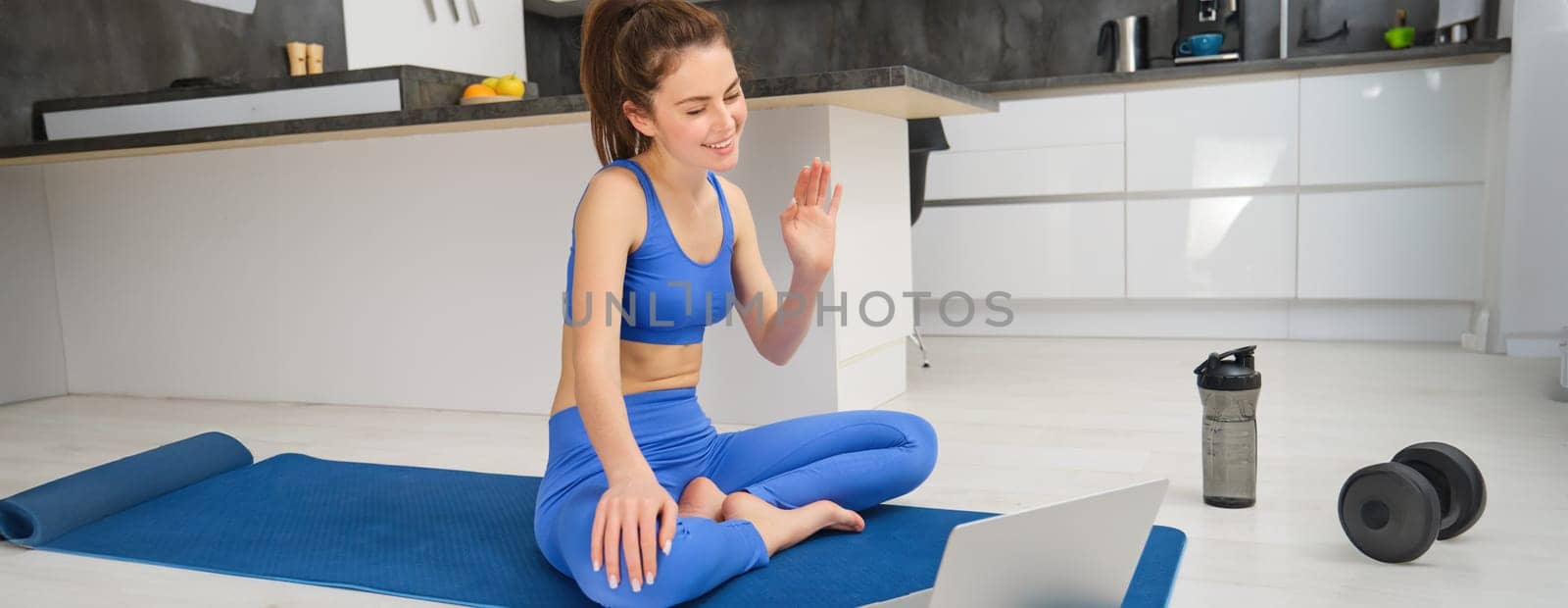 Portrait of fitness instructor, woman connects to online training session, waves hand at laptop, teaching yoga workout from home, sitting on rubber mat by Benzoix