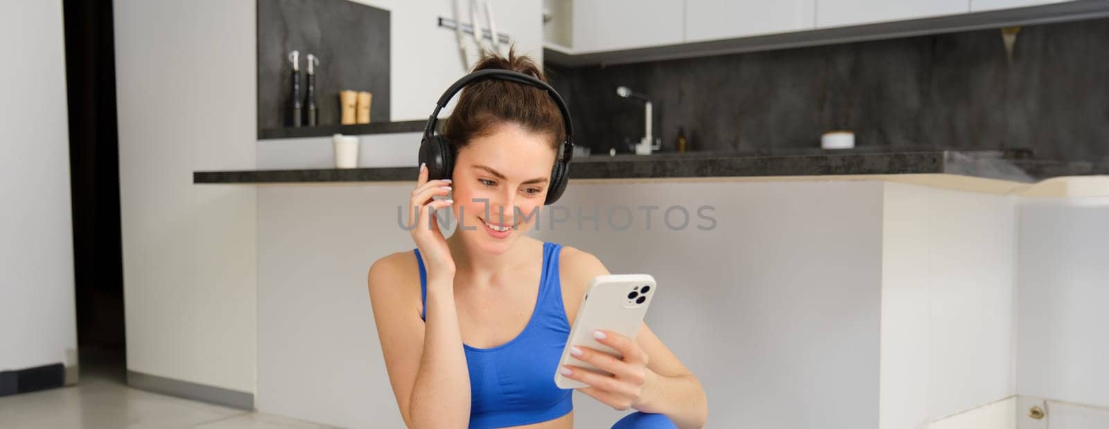 Portrait of smiling fitnes instructor, girl workout from home, wearing headphones and listens to music, holds smartphone.
