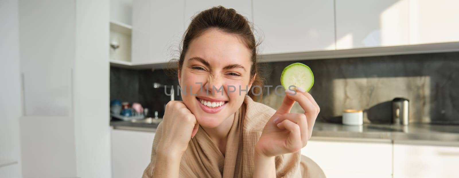 Portrait of beautiful brunette girl cooking in the kitchen, posing in bathrobe at home, holding zucchini, showing happy smile, making healthy food, vegetarian meal.