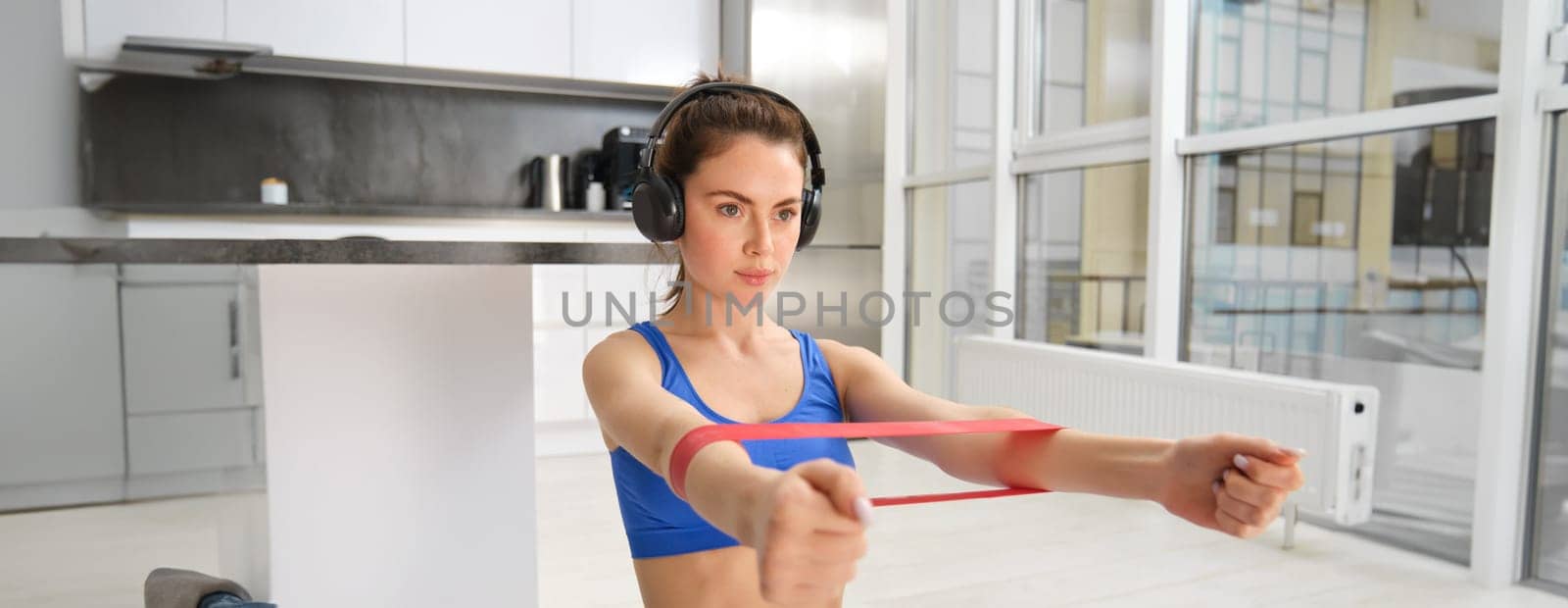 Image of hardworking sportswoman, girl doing fitness exercises at home, listening music in headphones, stretching elastic resistance band with arms, workout in living room by Benzoix