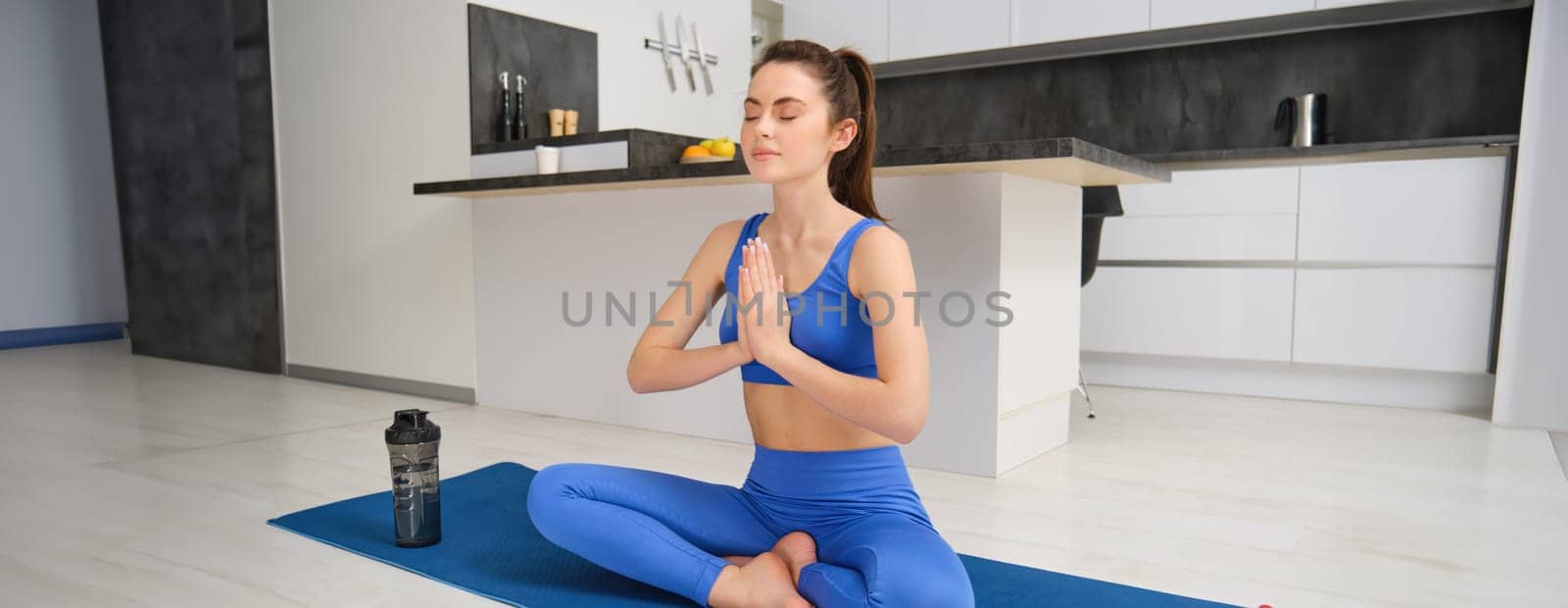 Sport and recreation. Close up portrait of beautiful girl does yoga at home, clasps her hands and meditates, sits on rubber mat in kitchen, makes asana, relaxing by Benzoix