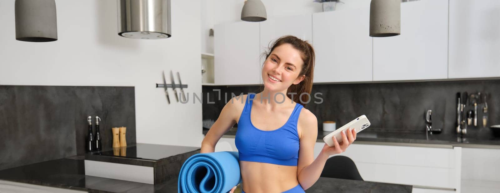 Image of strong and fit fitness instructor, woman standing at home with rubber mat and smartphone in hands, has an app for workout indoors by Benzoix