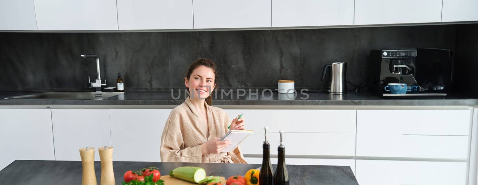 Portrait of woman thinking of menu, sitting in the kitchen and making grocery list, vegetables and meal ingredients on counter.