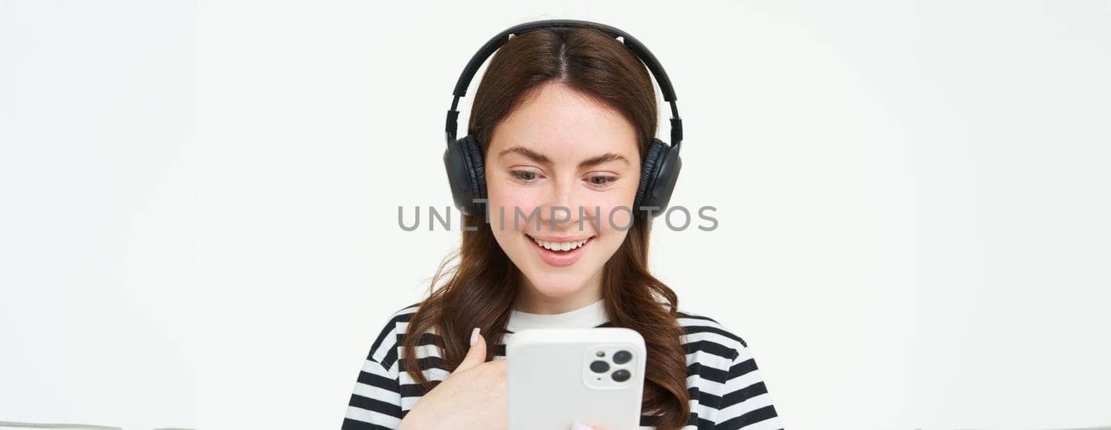 Image of young woman in headphones, using smartphone and laughing, watching video on mobile phone, listens to music on streaming service app, white background.