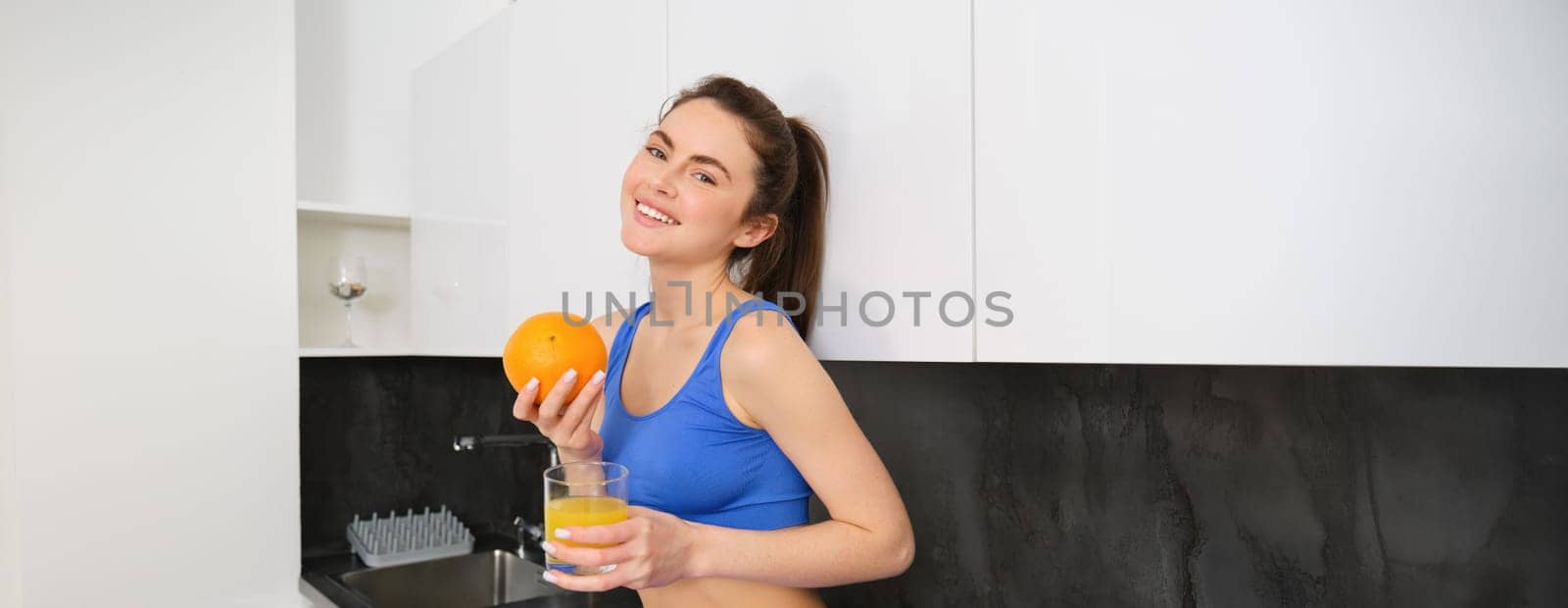 Indoor shot of woman after workout, standing in kitchen with fresh juice and an orange, drinking it by Benzoix