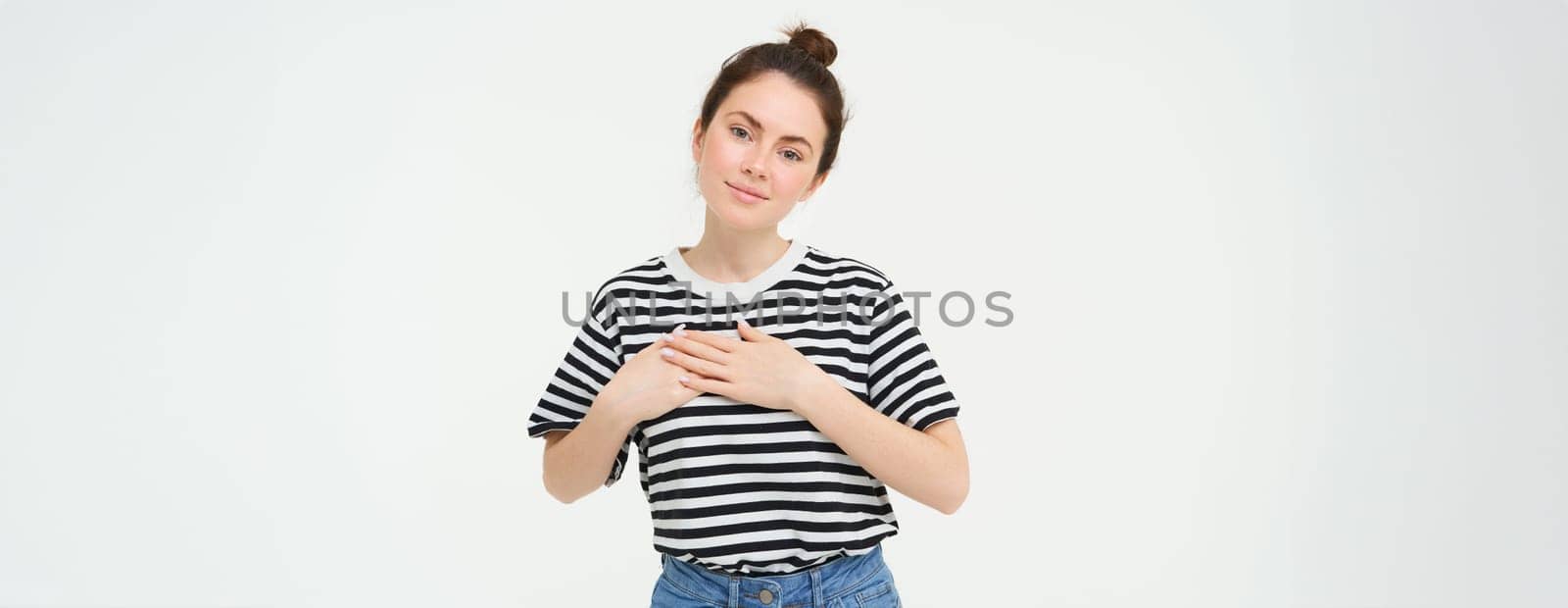 Portrait of happy, beautiful brunette woman, holds hands on heart and smiles, stands in casual clothes over white background. Copy space