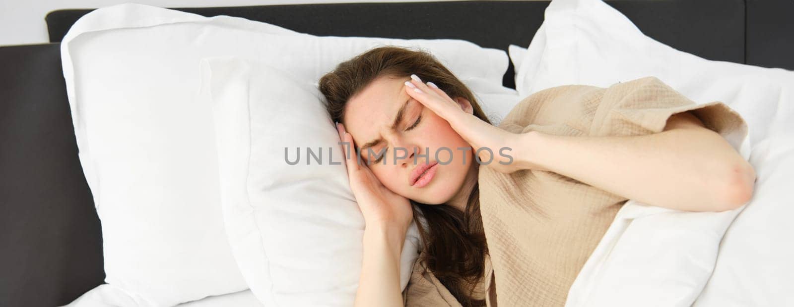 Woman lying in bed and feeling unwell, having headache, touching her head with frustrated face, has high fever or migraine. concept of health and people.
