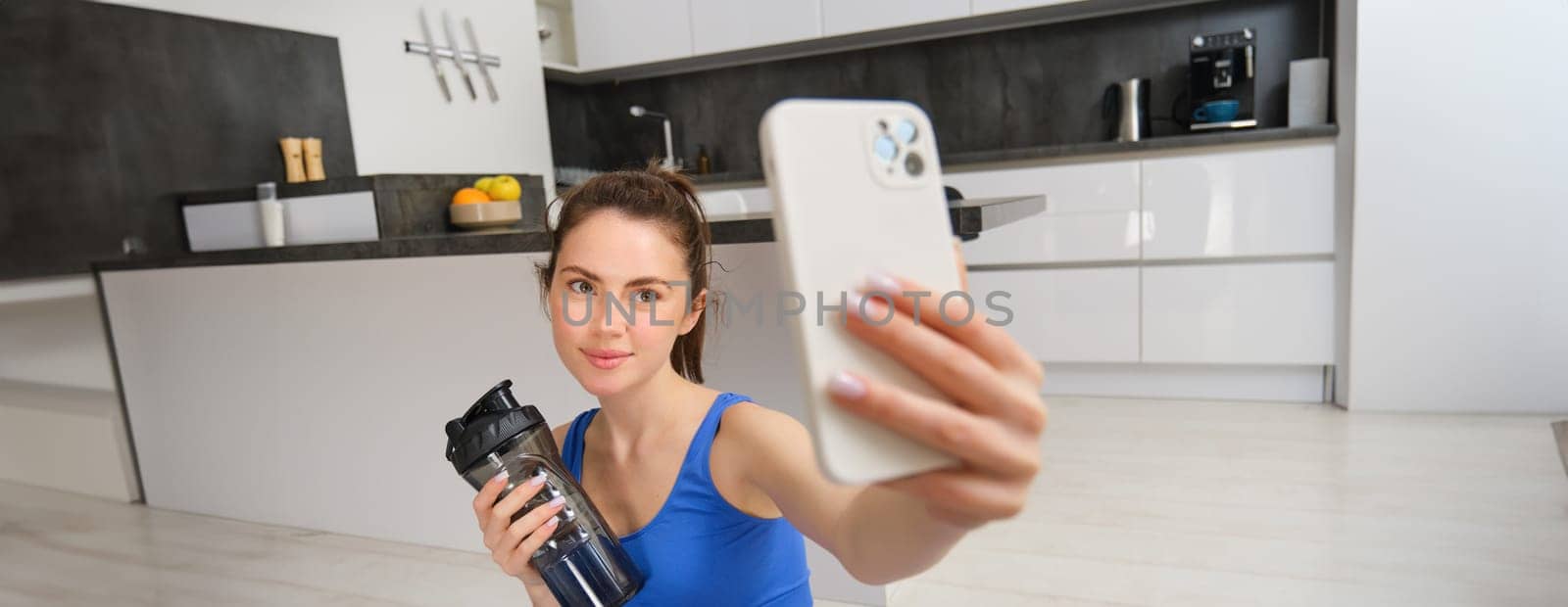 Portrait of young fitness girl, takes selfie on smartphone with water bottle, drinks water during workout, training at home, records sports vlog by Benzoix