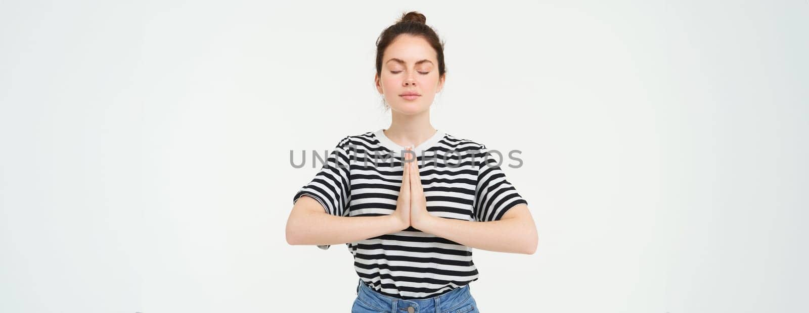 Portrait of young mindful woman meditating, holds hands clasped together, namaste gesture, practice yoga, standing over white background by Benzoix