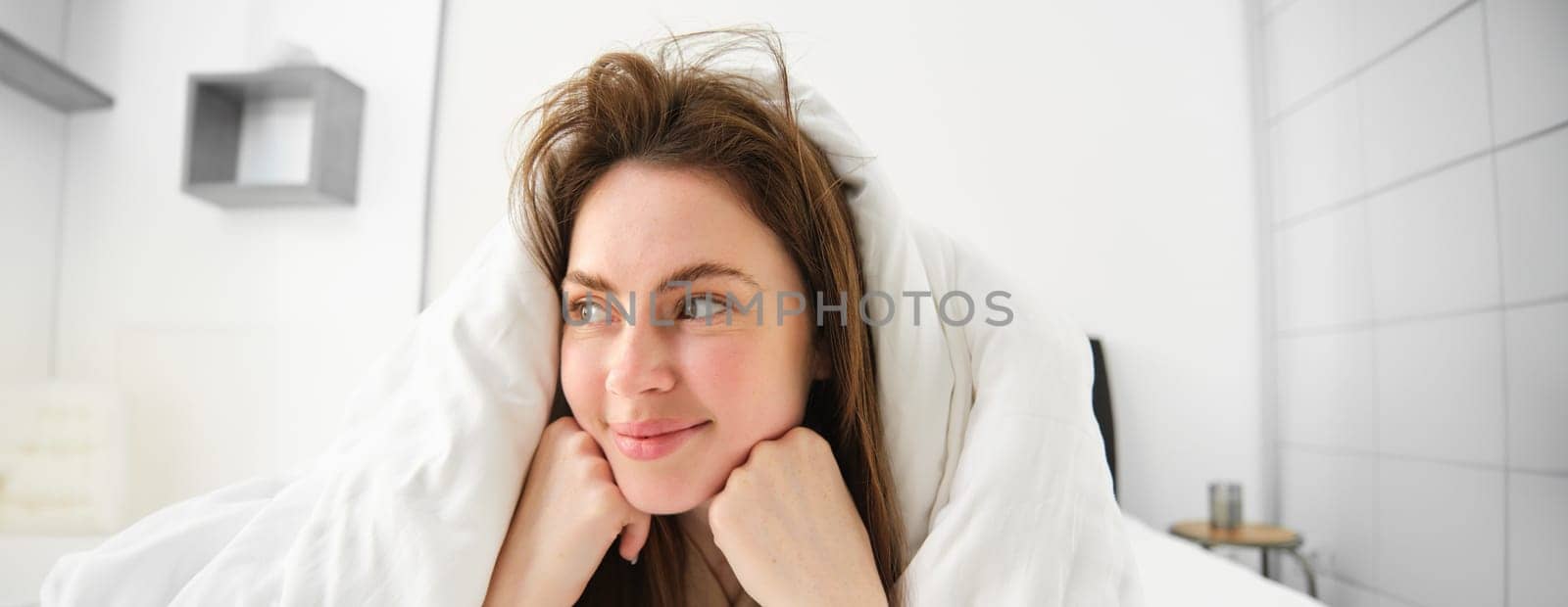 Cute girl with messy hair, lying in bed covered in white sheets duvet, smiling and laughing coquettish, spending time in her bedroom by Benzoix