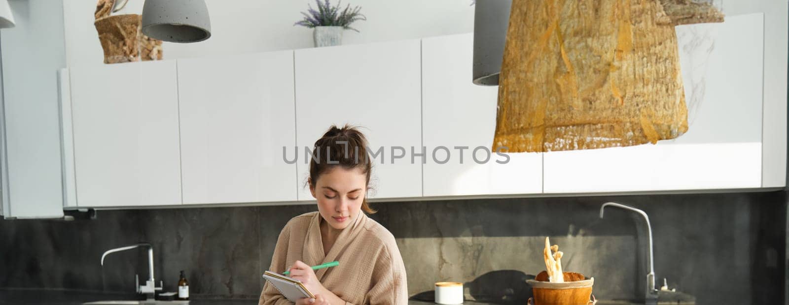 Vertical shot of young woman writing down food ideas, creating new meal, writing down grocery list or recipe in the notebook, standing in the kitchen near chopping board with fresh vegetables by Benzoix