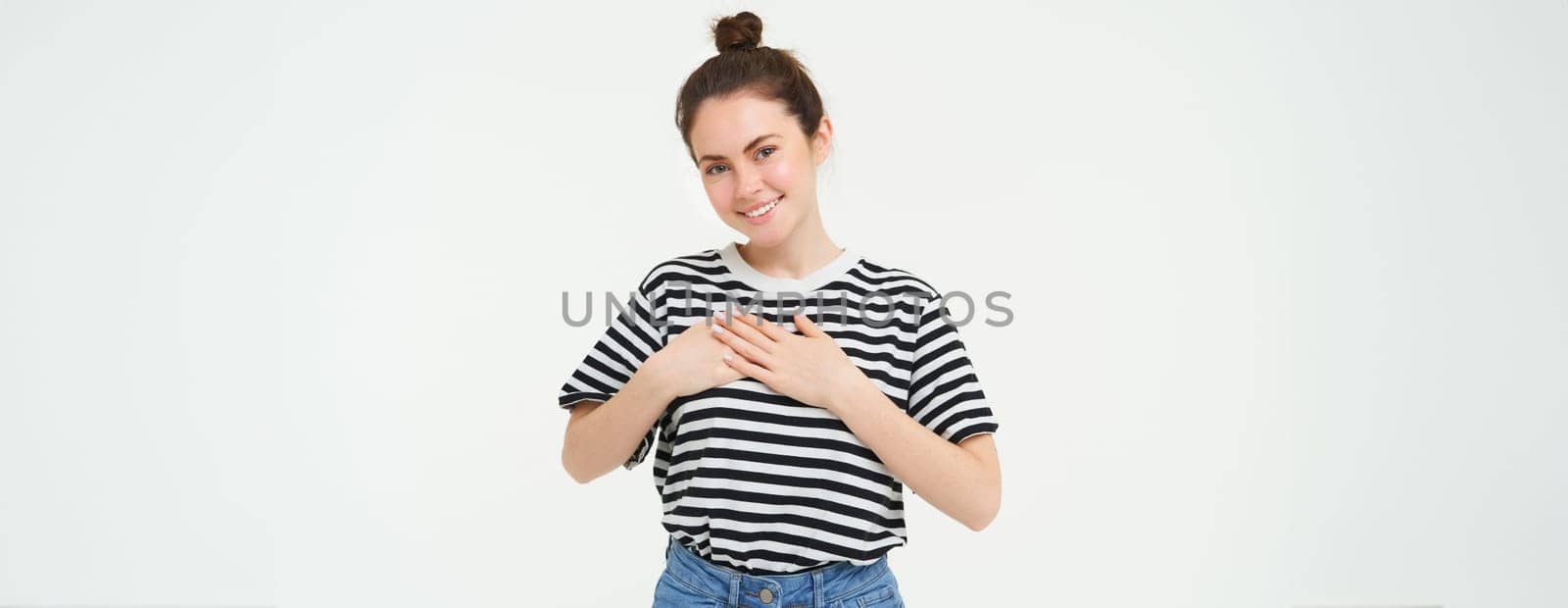 Young smiling woman, feels gratitude, thankful for something, holds hands on heart and looks tender at camera, standing over white background.