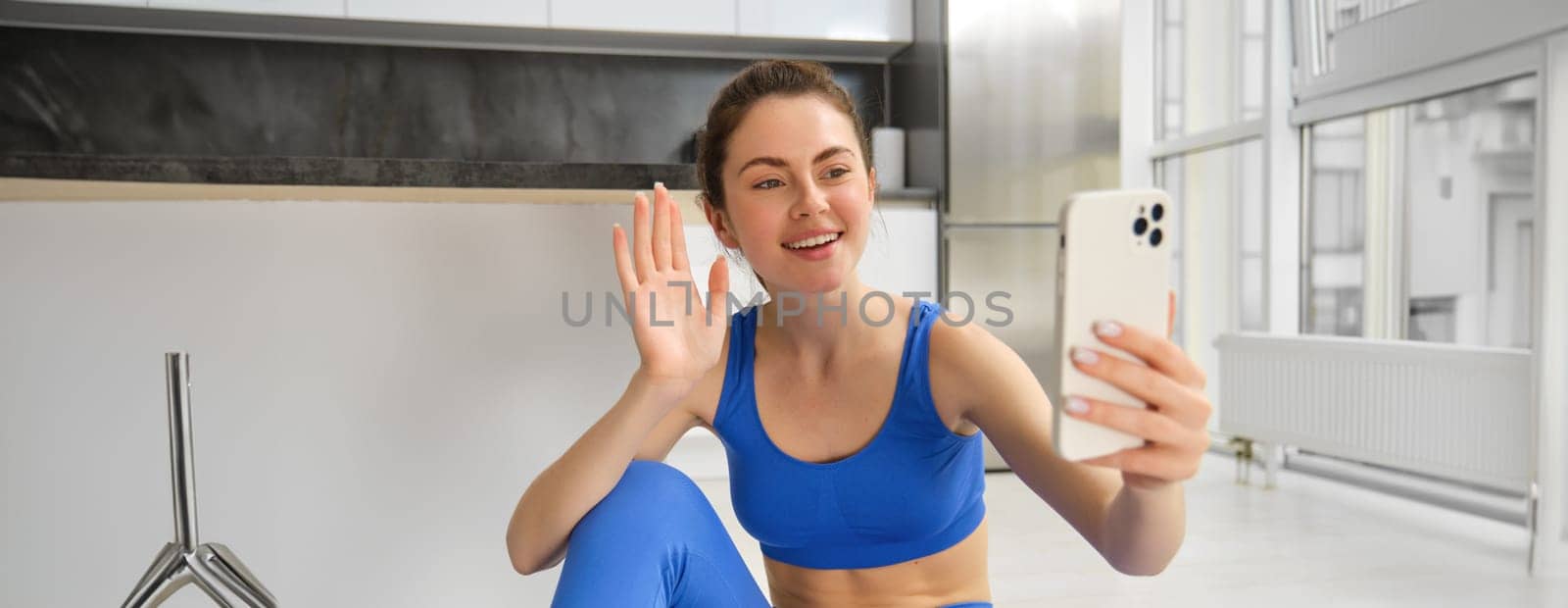 Image of young woman doing workout, takes selfie, records online video. Fitness instructor records her training on phone, says hello at smartphone camera and waves hand at it.