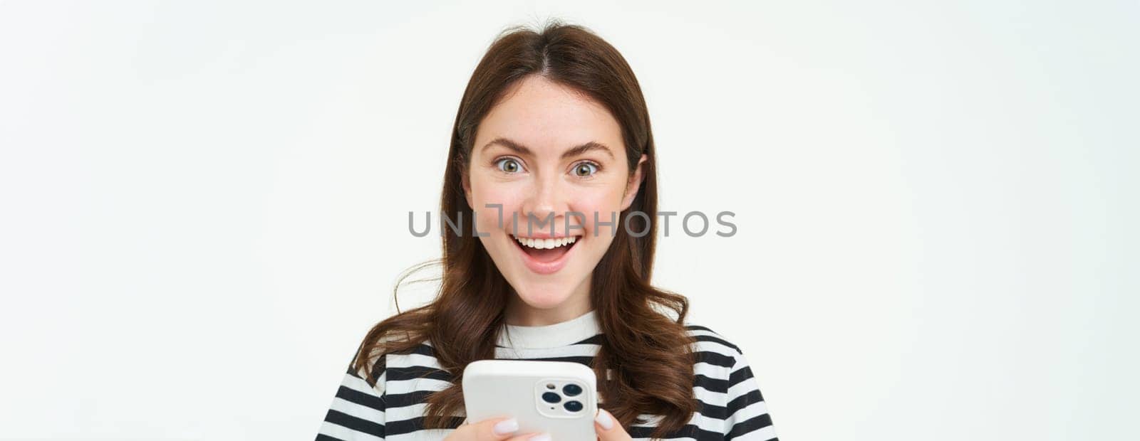 Portrait of excited brunette girl, reacting to amazing promo on smartphone, holding mobile phone and smiling, isolated on white background.