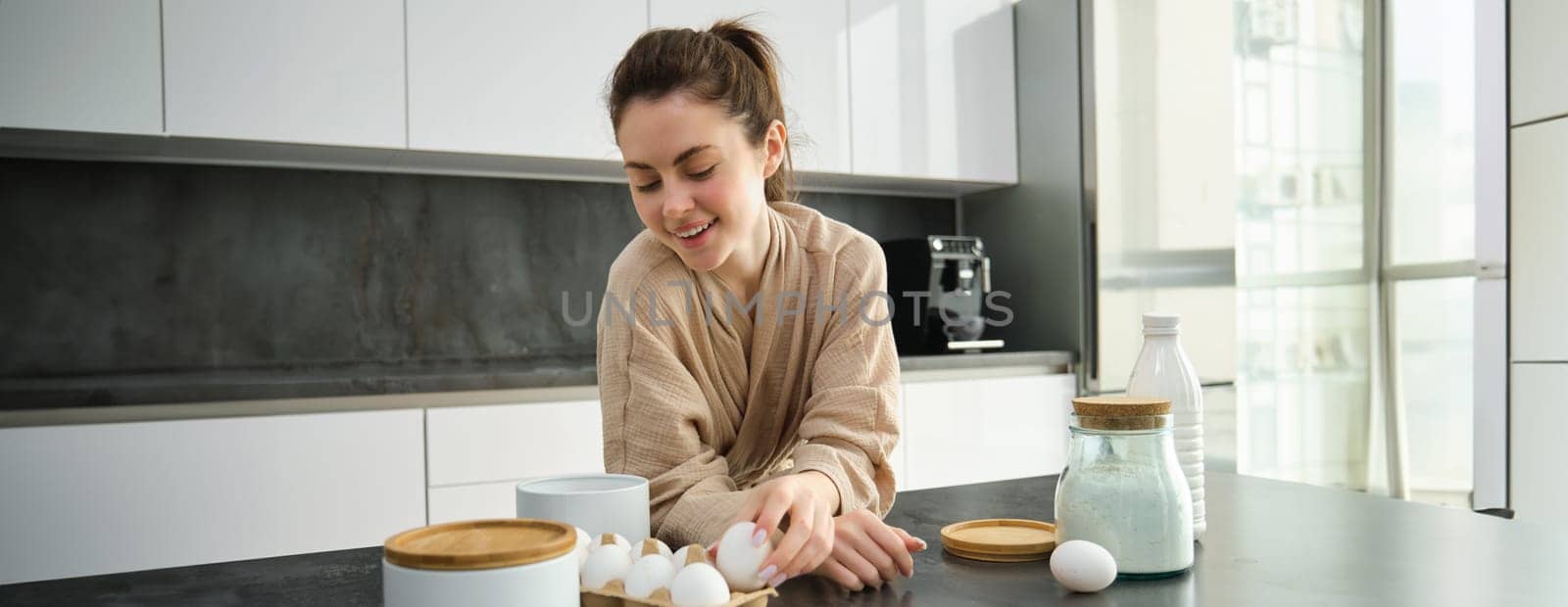 Attractive young cheerful girl baking at the kitchen, making dough, holding recipe book, having ideas.