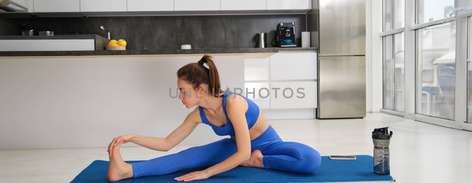 Image of young fit woman, does gymanstics at home, stretches her legs on floor in living room, does fitness workout, yoga warm-up exercises.