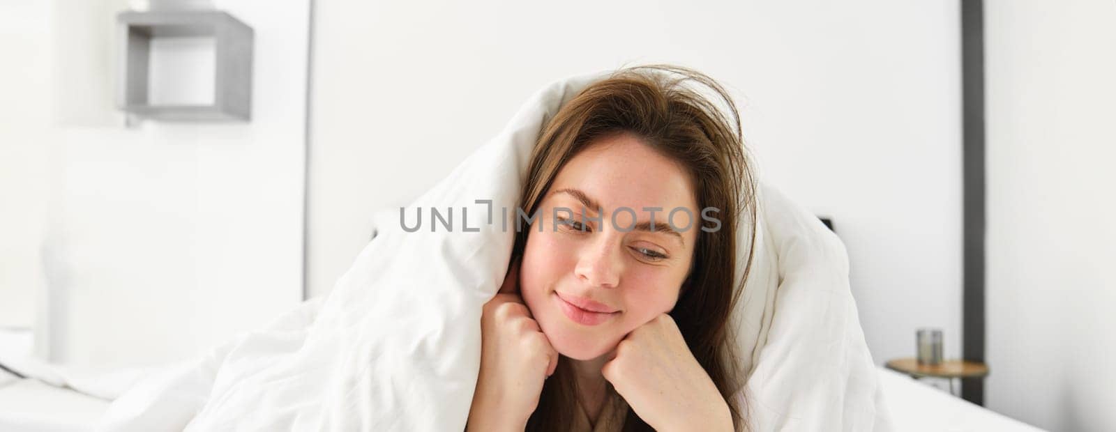 Cute girl with messy hair, lying in bed covered in white sheets duvet, smiling and laughing coquettish, spending time in her bedroom.