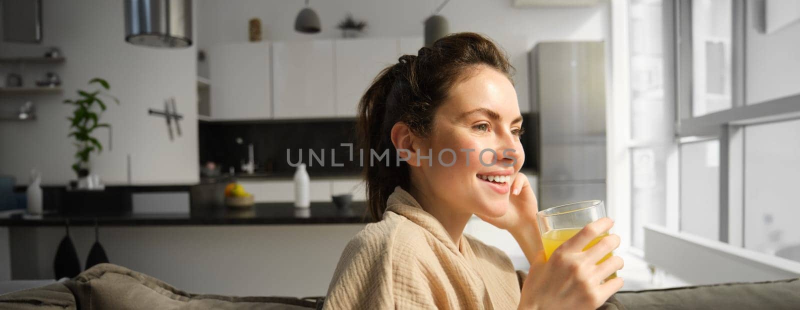 Close up of beautiful young woman on sofa, drinks orange juice and smiles, looks outside window, enjoys morning at home.
