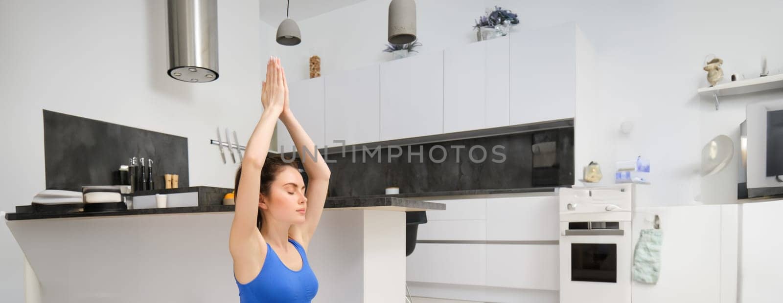 Vertical shot of young beautiful woman doing fitness training, lifts her hands up and breathing exercises, meditating at home on rubber mat.