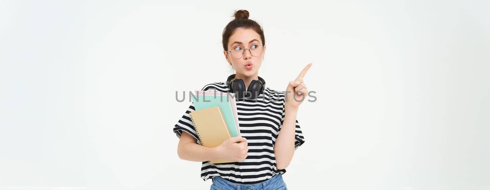 Image of intrigued girl in glasses points and looks left at smth interesting, holds documents, white background.