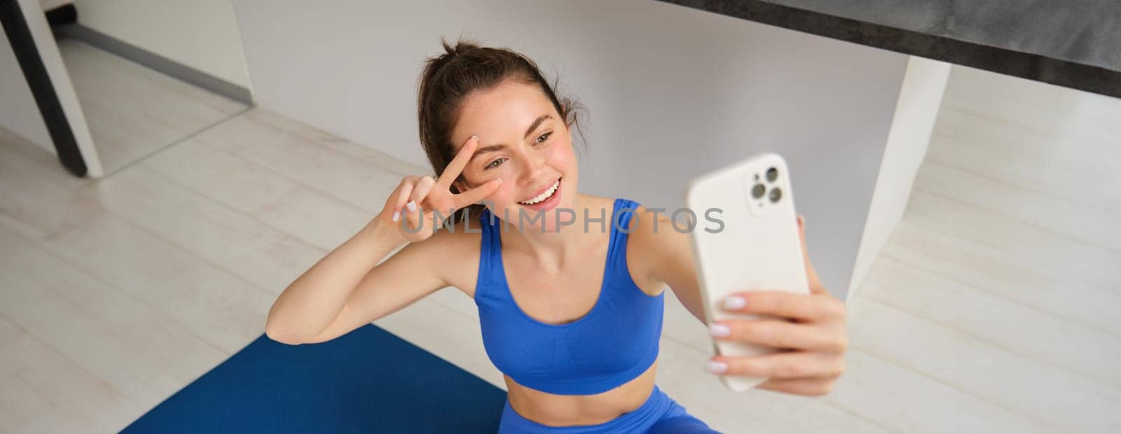 Portrait of beautiful female athlete, woman doing workout at home on rubber yoga mat, takes selfie on smartphone, makes photos for social media of fitness instructor.