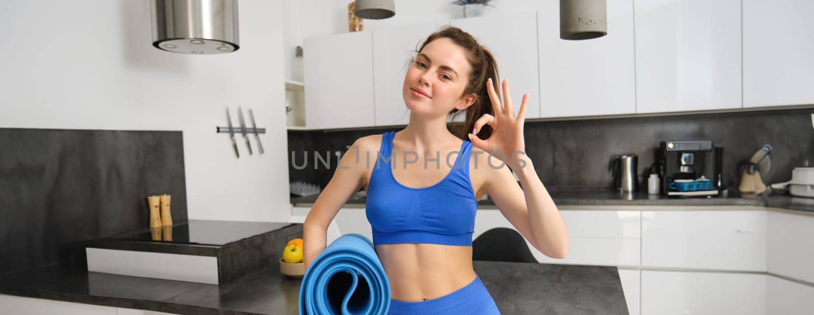 Smiling brunette woman, fitness instructor approves yoga app, mindfulness application, workout at home, standing in living room, showing ok gesture and holding rubber mat by Benzoix