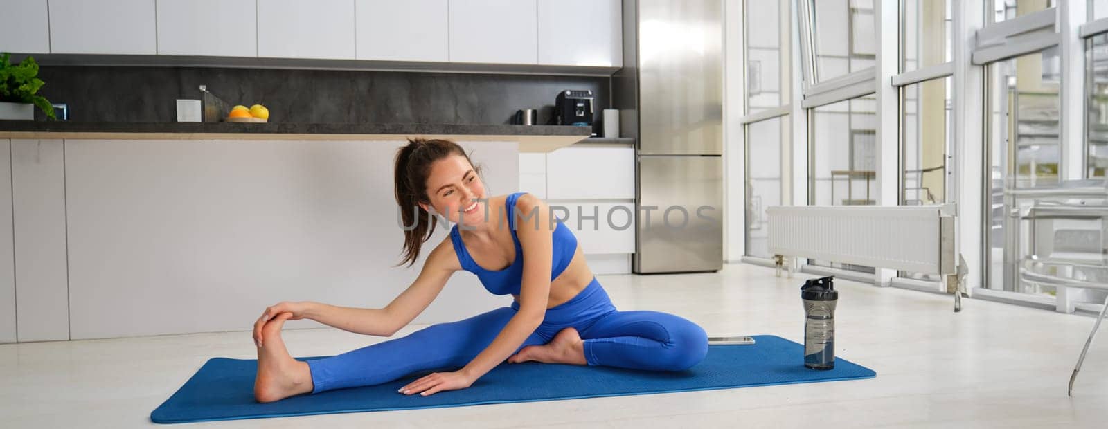 Portrait of young fitness woman doing aerobics exercises, gymnastics training at home, stretching her legs before splits workout, sitting on floor, rubber blue mat.