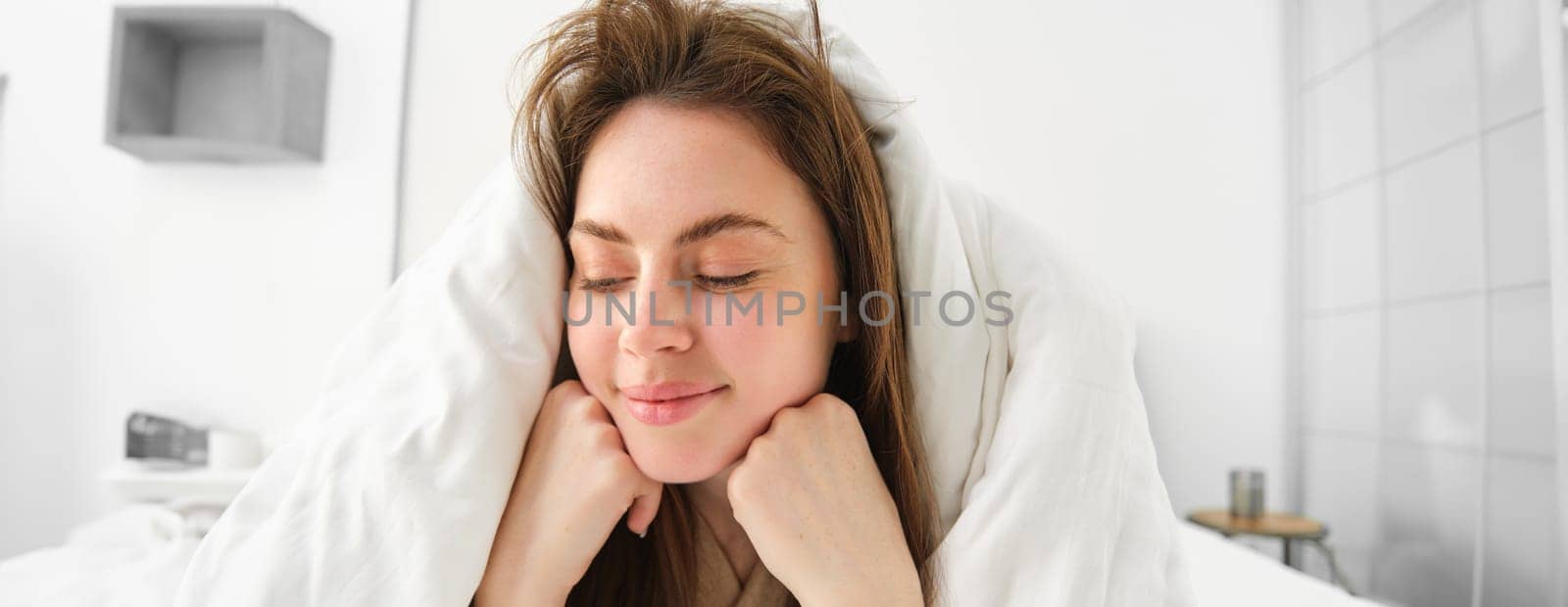 Cheerful woman feeling comfortable in bed, lying in bedroom covered in white sheets, smiling pleased, has messy hair in morning, waking up and looking happy.