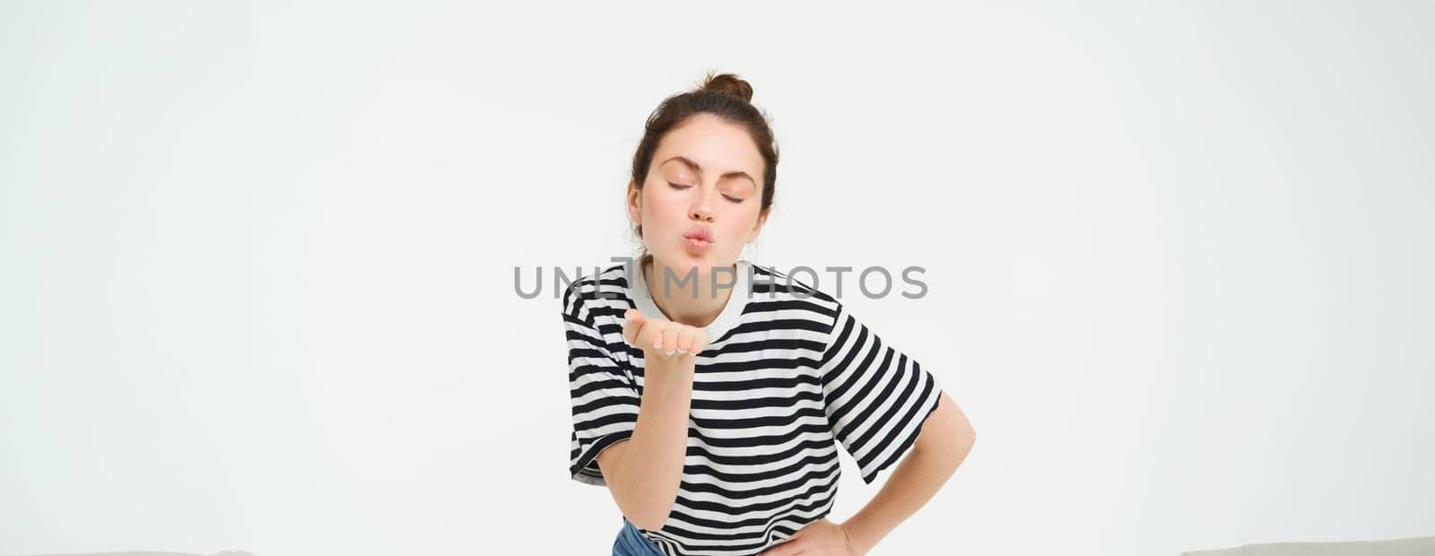 Portrait of beautiful brunette woman sends air kiss at camera, holds palm near lips, isolated against white background.