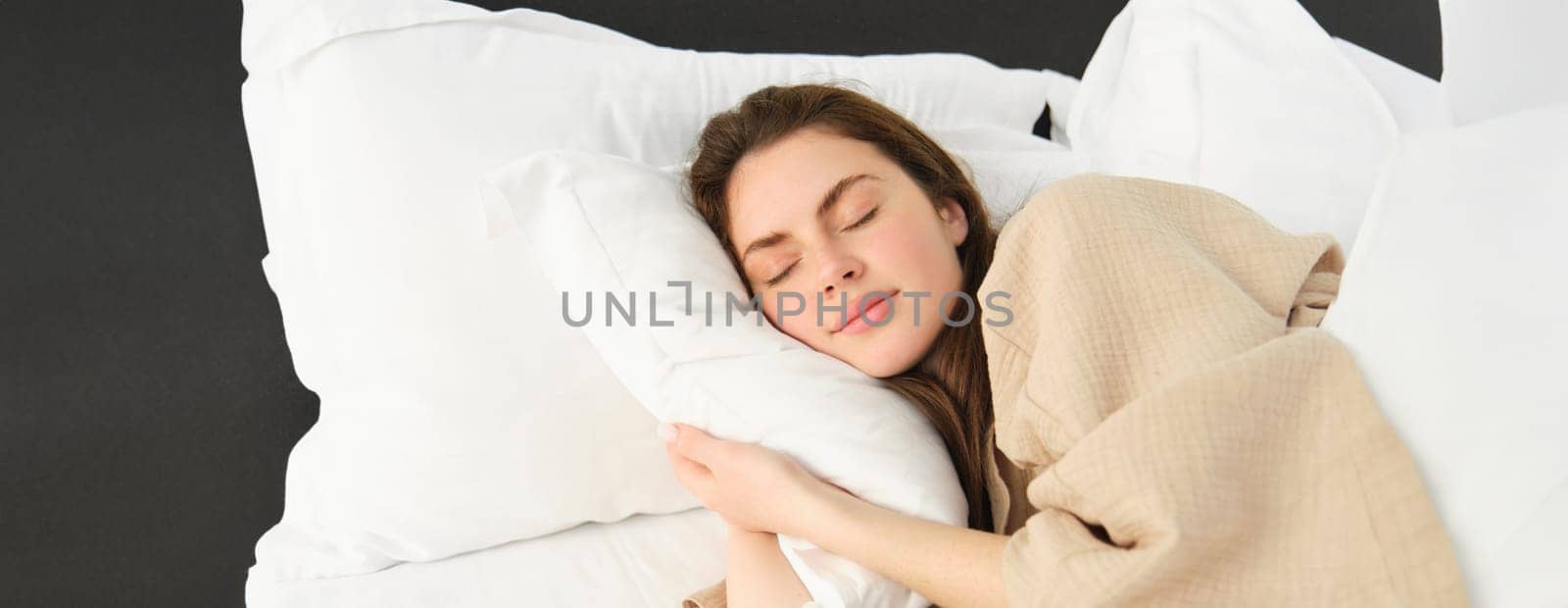 Portrait of happy, beautiful young woman resting in bed, wearing pyjamas, hugging her pillow and smiling while sleeping.