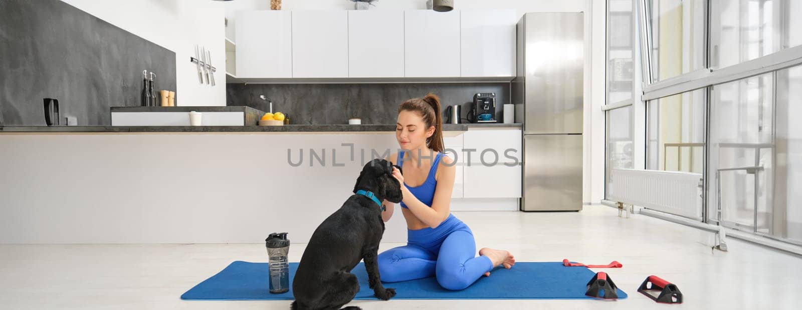 Image of young woman does workout from home, sits on yoga mat in living room and plays with her black dog, puppy distracts girl from doing exercises.
