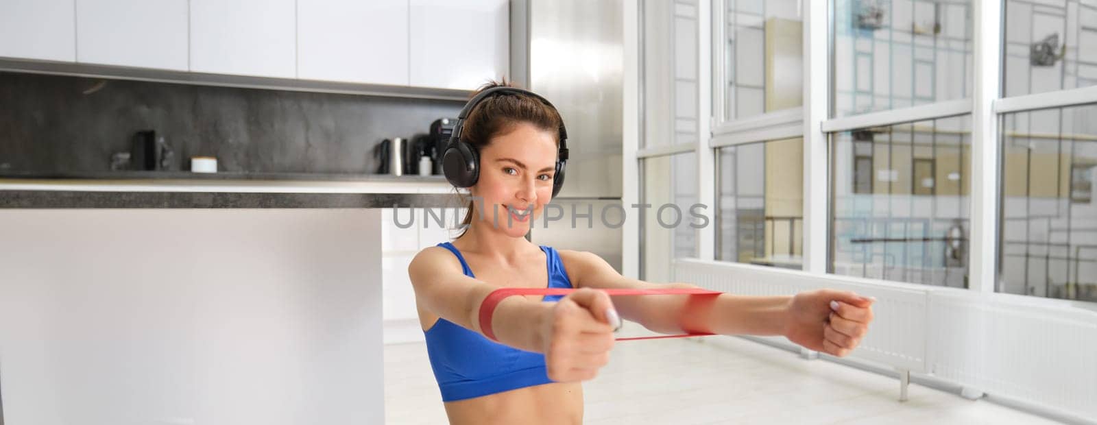 Focused sportswoman workout at home, using elastic resistance band on arms, stretching exercises in living room, aerobics training.