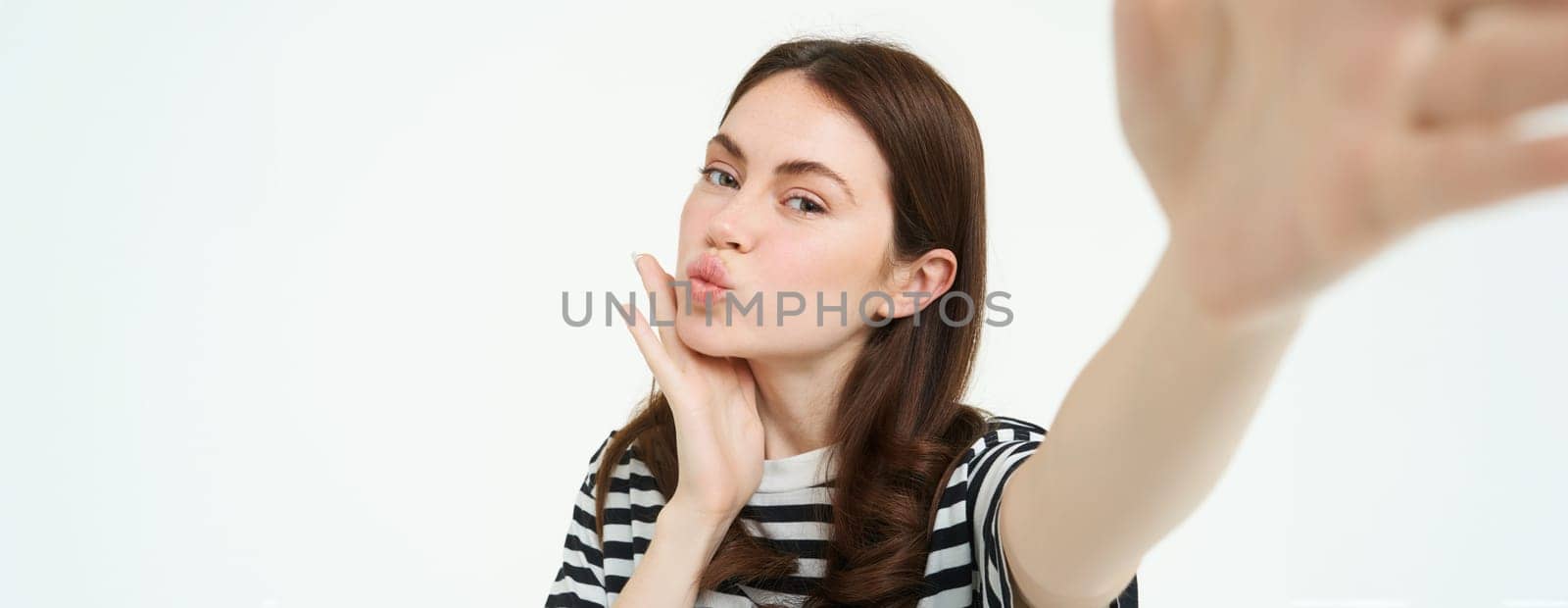 Portrait of cute, feminine girl posing near something, taking selfie on smartphone, holding camera with extended hand, isolated on white background.