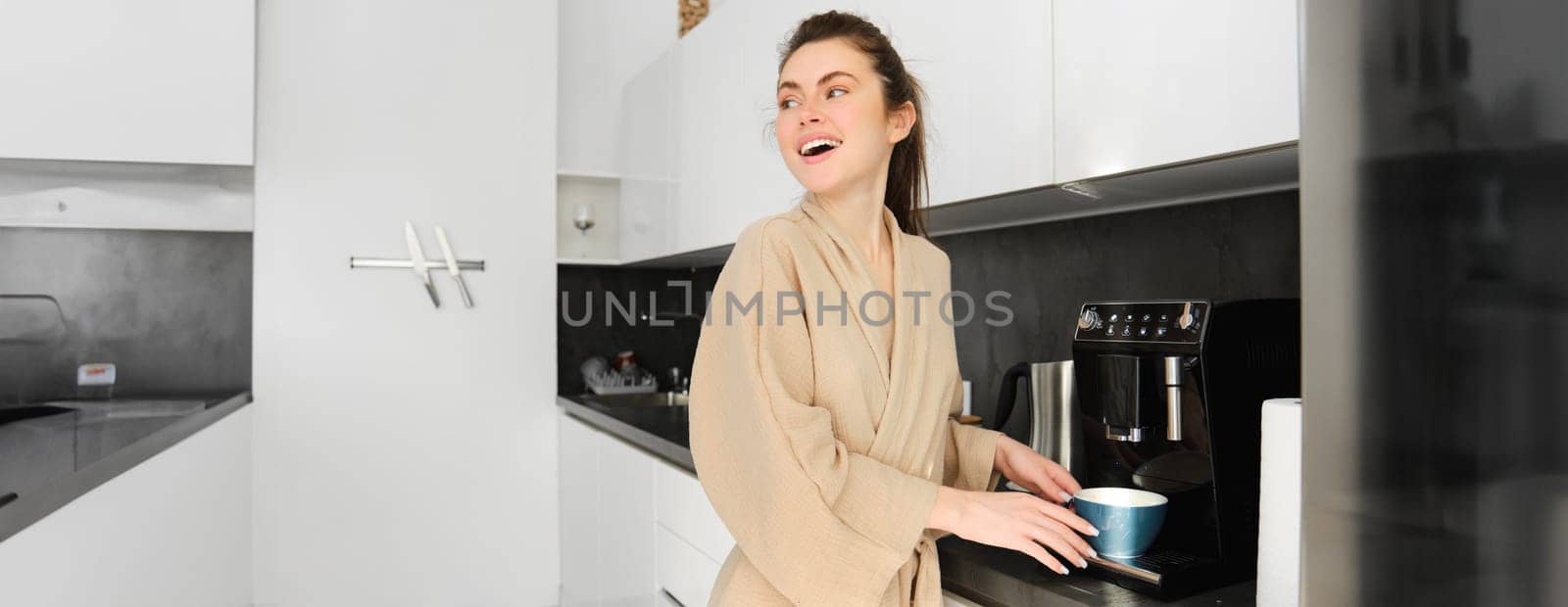 Portrait of happy smiling woman in the kitchen, using coffee machine to make herself morning mug, preparing cappuccino, wearing bathrobe.