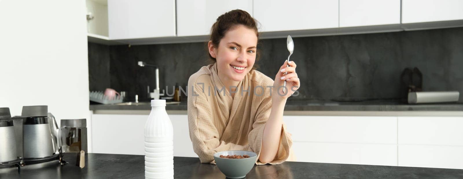 Portrait of beautiful young and healthy woman in bathrobe eats her breakfast in kitchen, has cereals with milk and smiling.